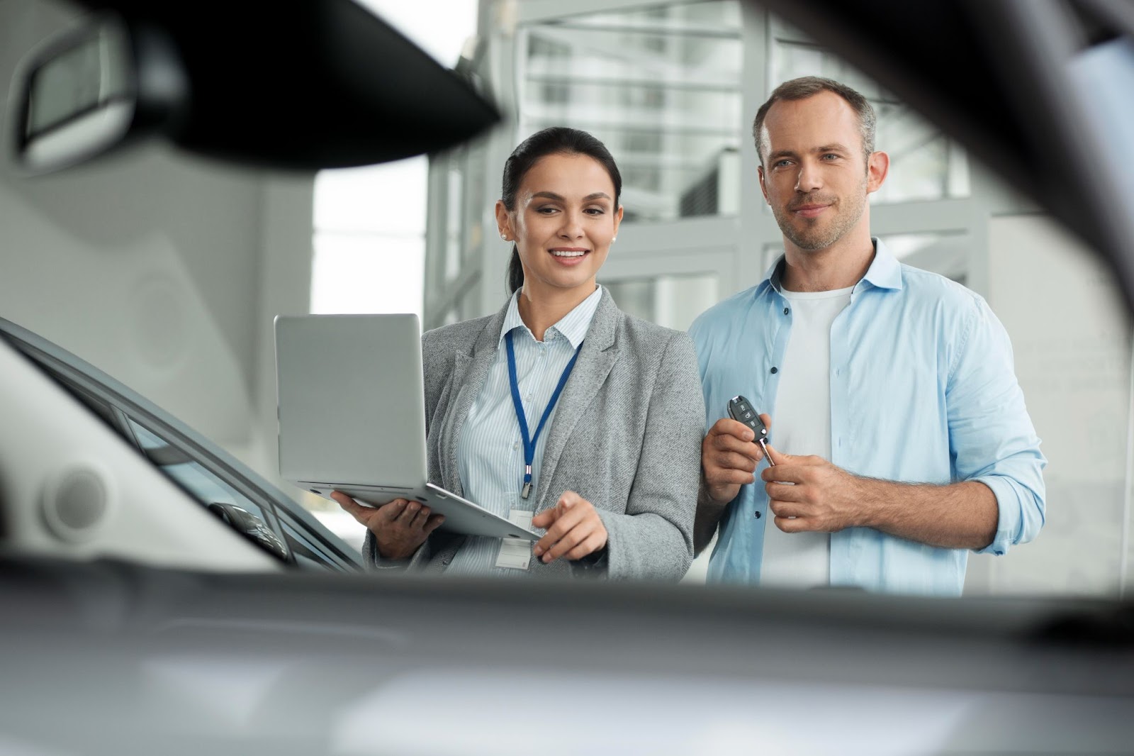 Uma mulher de blazer e crachá aponta para um carro, enquanto segura um notebook aberto na ficha técnica de carros, ao lado dela um homem de camisa azul segura a chave do veículo.