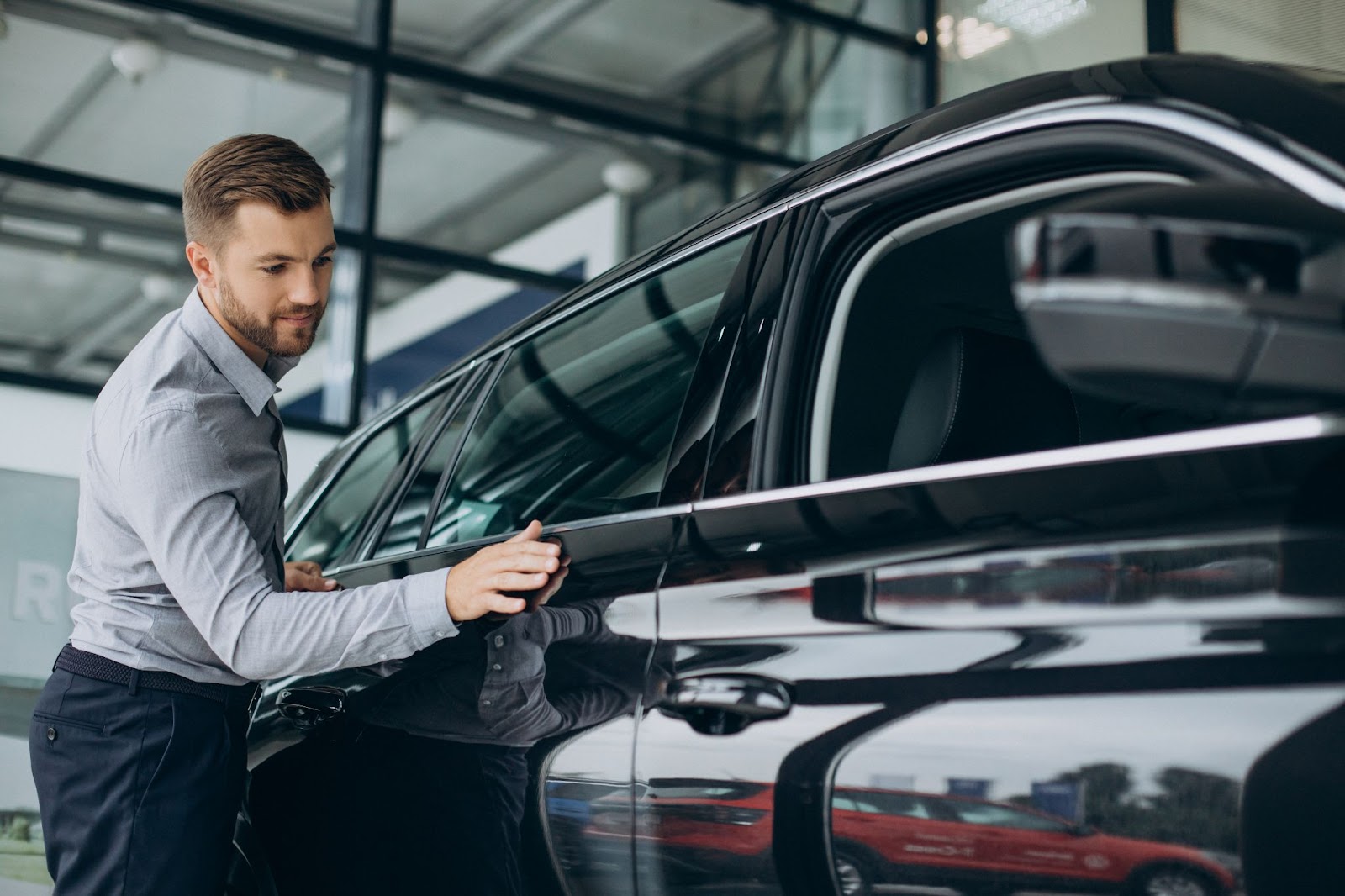 Um homem de camisa social cinza está ao lado de um carro preto e o observa enquanto passa a mão delicadamente pela carroceria.