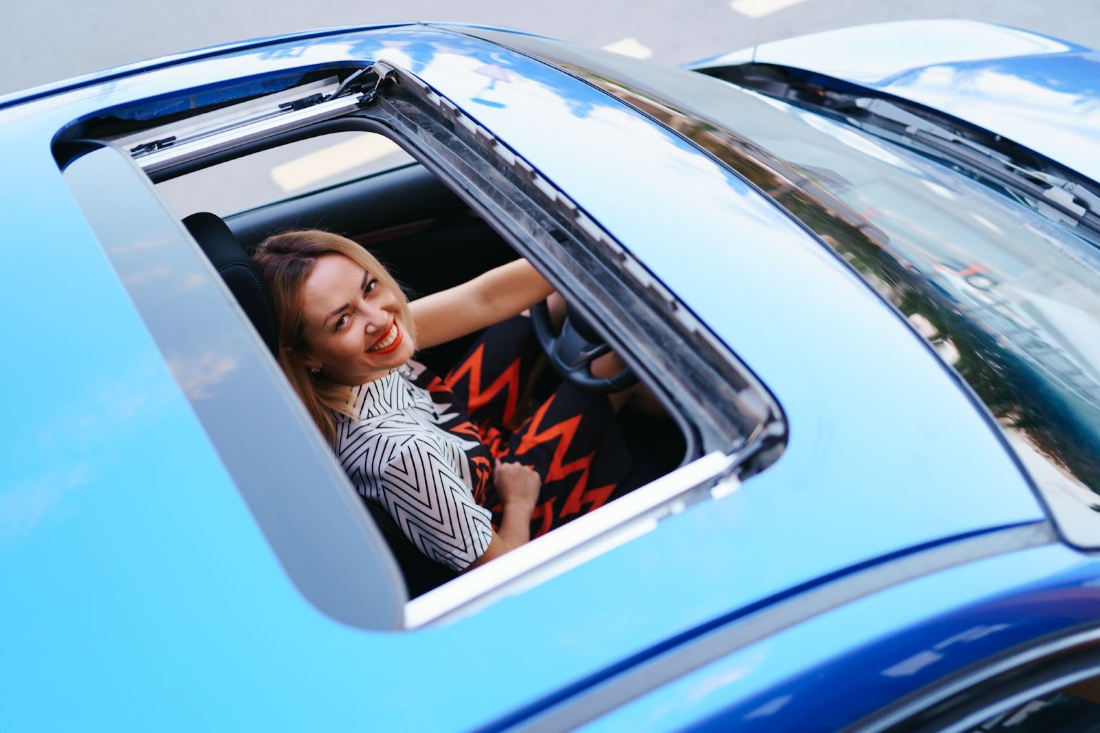 Visão superior de um carro azul, com o teto solar aberto, revelando a motorista loira sorridente.