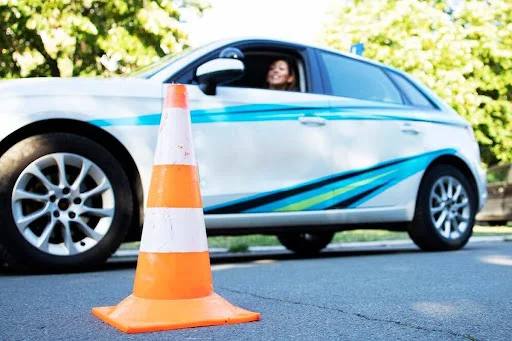 A foto mostra uma mulher conduzindo um veículo e olhando para um cone posicionado na rua.