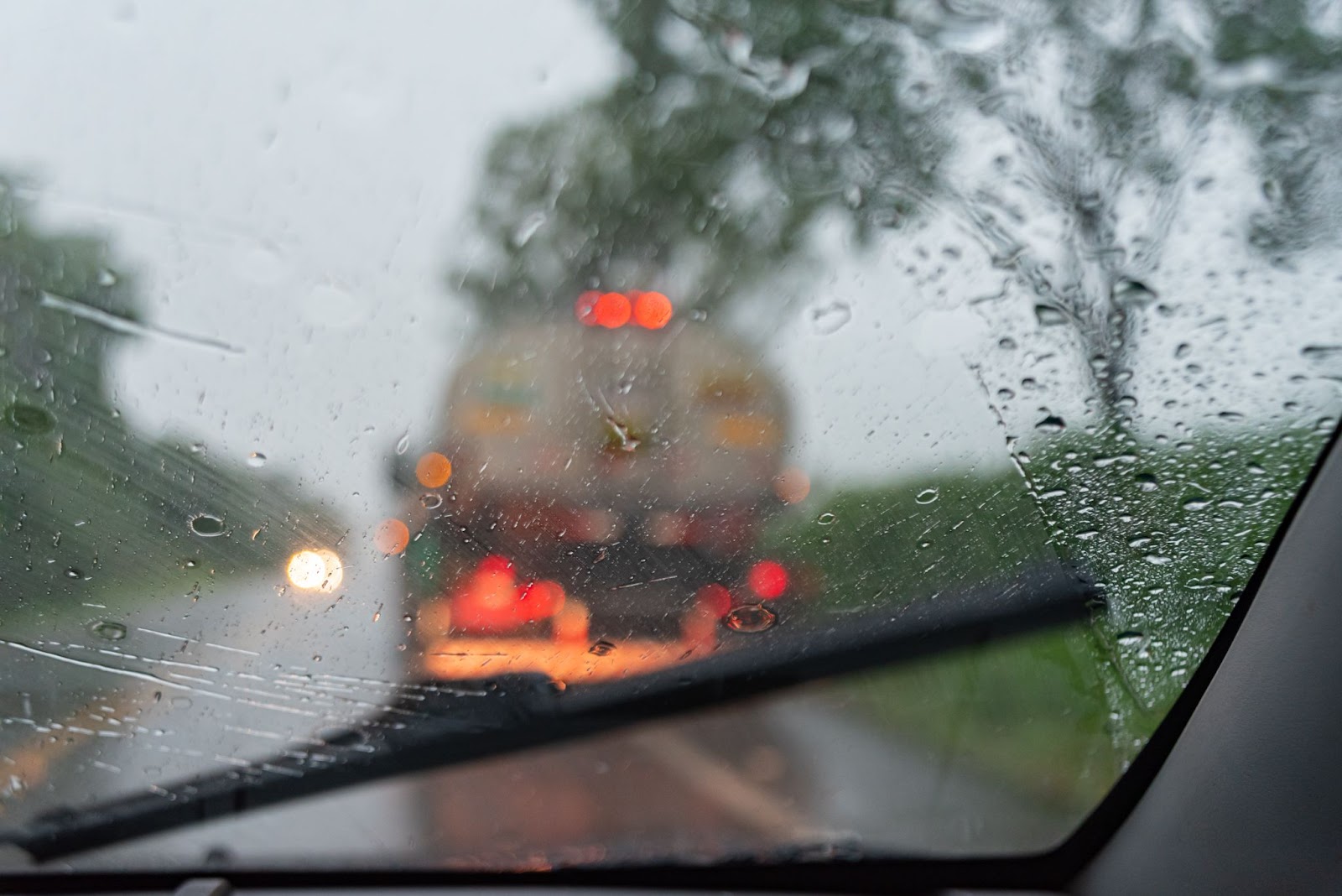 A imagem em close-up mostra a visão de dentro de um carro, com o vidro da frente embaçado por dirigir na chuva. Na frente do carro, é possível ver um caminhão na estrada desfocado.