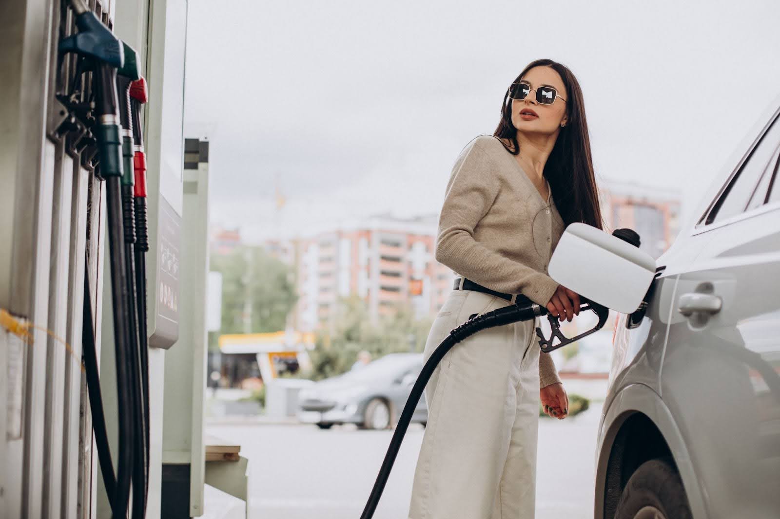 Na imagem uma mulher de óculos escuros abastece seu carro enquanto observa a bomba de gasolina.