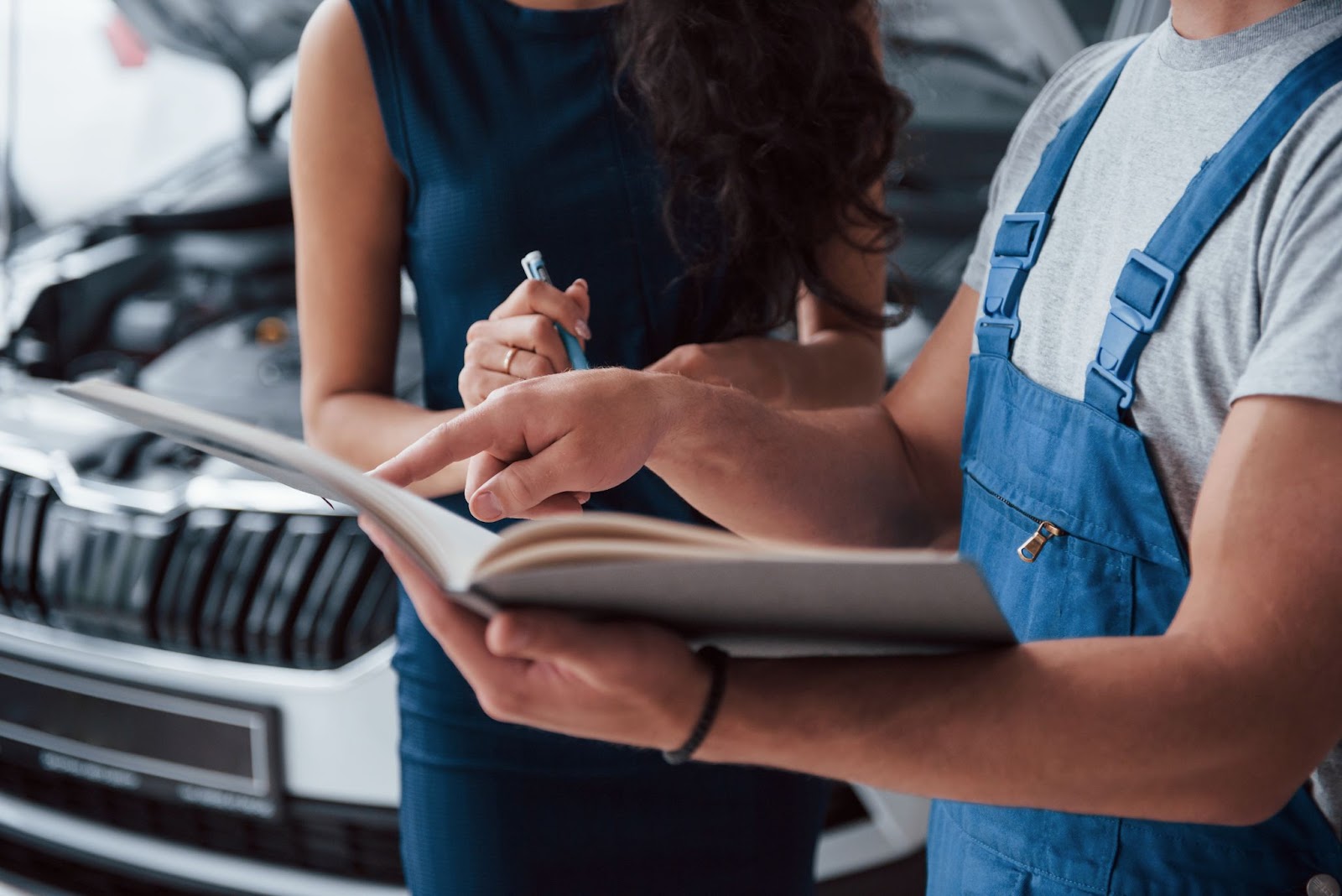 Um homem de macacão aponta para uma ficha técnica de carros, ao lado dele há uma mulher de vestido azul, segura uma caneta.