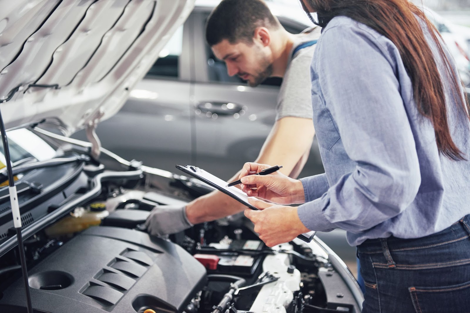 Na imagem, há uma mulher com uma prancheta na mão, supervisionando um profissional de luvas que avalia se o motor de um carro irá receber o certificado de segurança veicular.