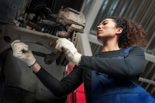 Uma mulher mecânica aparece na imagem usando macacão e luvas, inspecionando bucha da bandeja na parte de baixo de um veículo.