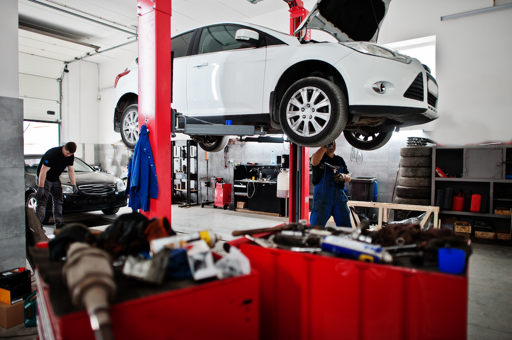 Carro branco suspenso em uma oficina mecânica onde um profissional realiza reparos, ao fundo outro mecânico verifica um carro preto.