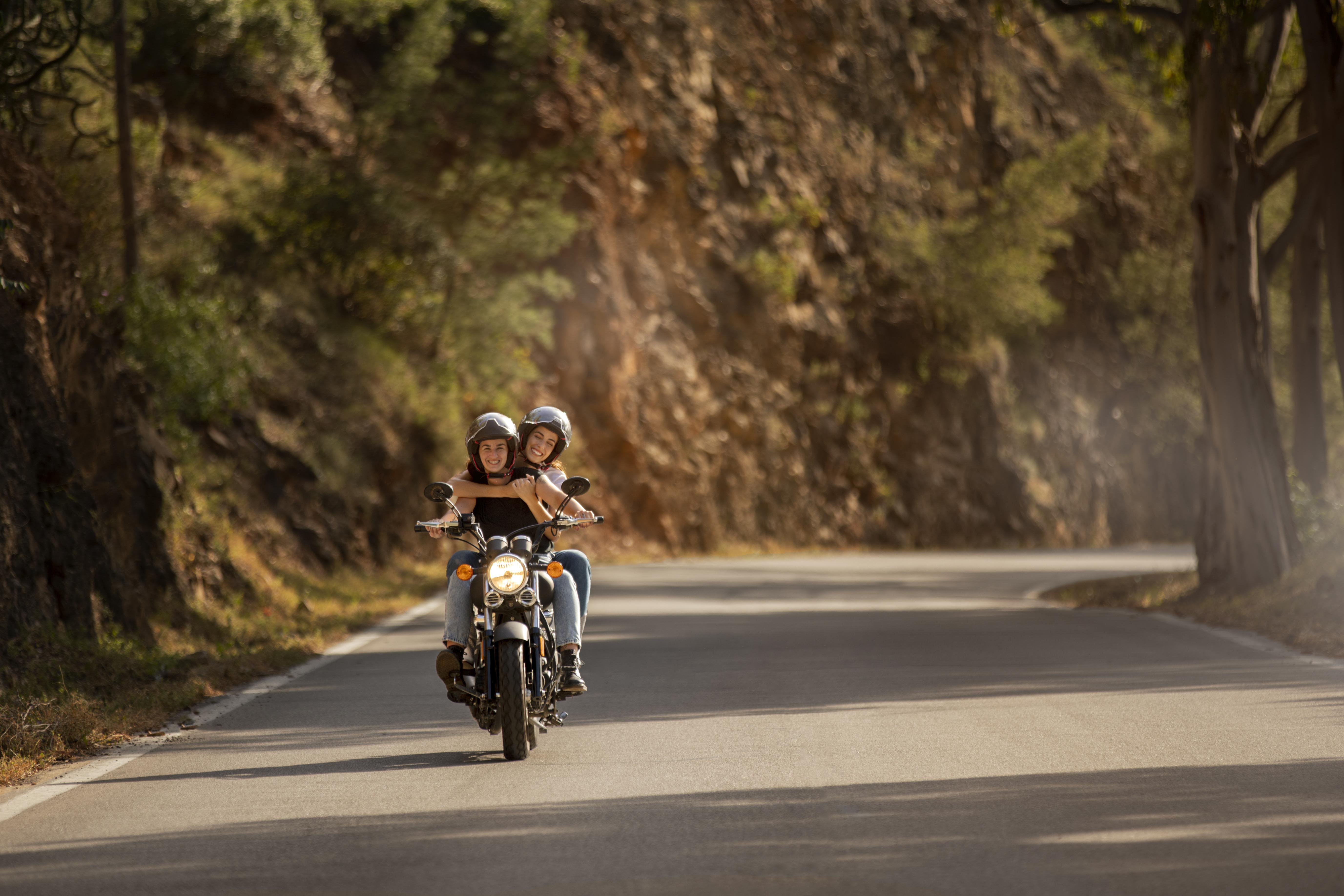 Imagem de duas mulheres que sabem qual a melhor bateria de moto andando em uma motocicleta em uma estrada durante o dia. Elas vestem capacete e têm expressão feliz.