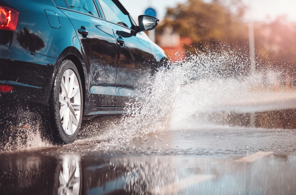 Close-up de um carro em movimento, dirigindo na chuva e passando por uma poça d'água.