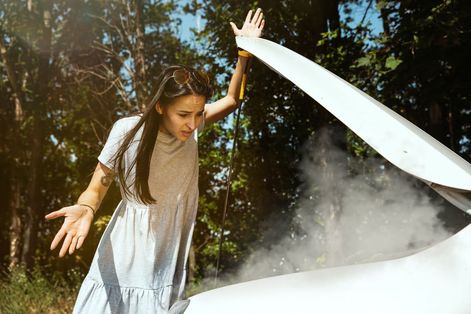 Na imagem, uma mulher de vestido cinza está em frente a um carro branco com o capô aberto de onde sai fumaça, representando uma das consequências de um sistema de arrefecimento defeituoso.