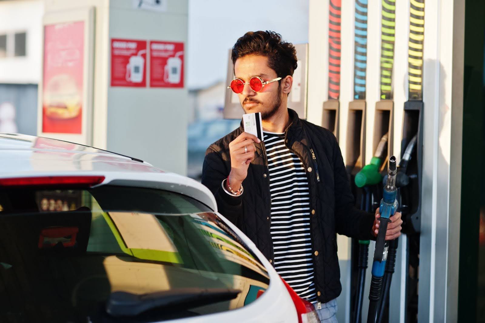 Na imagem, um homem de jaqueta preta está parado em um posto de gasolina, ao lado de um carro. Ele parece pensativo, tentando tomar a decisão entre abastecer o veículo com gasolina aditivada ou comum.