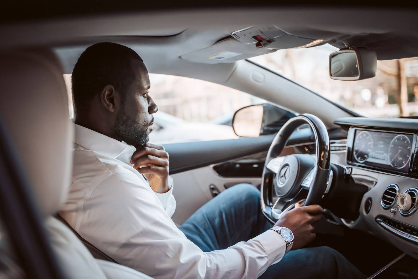 Homem de camisa social branca, sentado no banco do motorista, com uma expressão pensativa, apoiando a mão direita no volante e a esquerda no queixo.