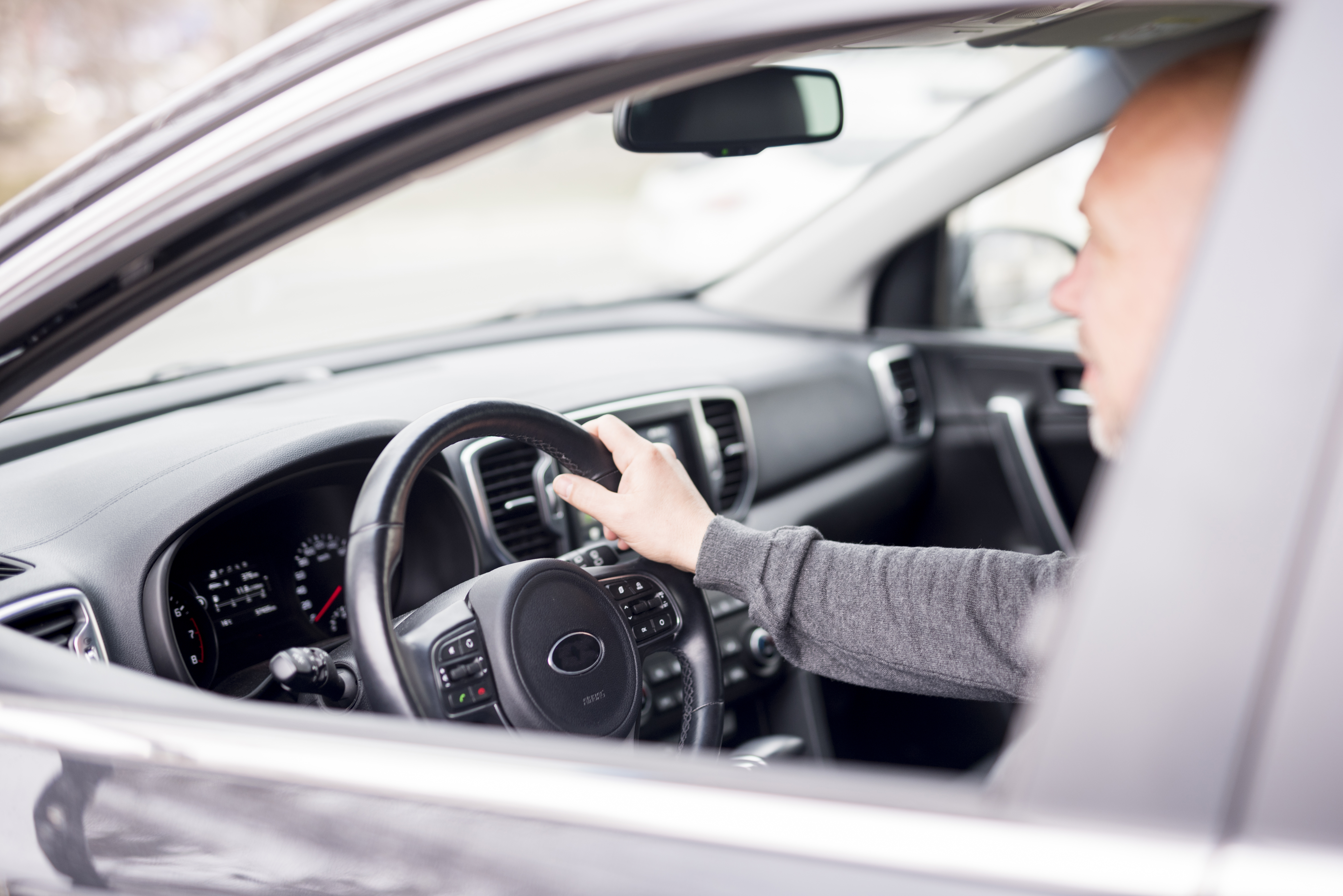Imagem de fora do carro mostra homem idoso de blusa cinza, dirigindo com a mão direita no volante de direção hidráulica.