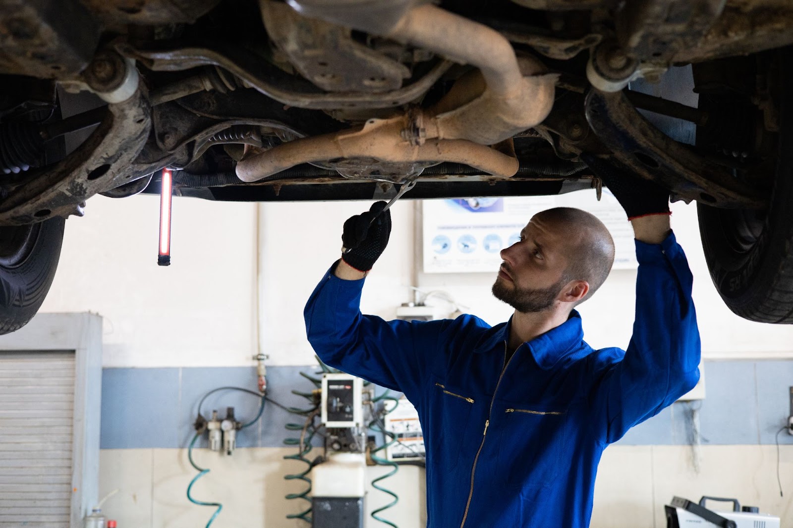 Em uma oficina, um mecânico vestindo um macacão azul realiza a manutenção preventiva em um veículo, verificando as peças inferiores com uma ferramenta apropriada.