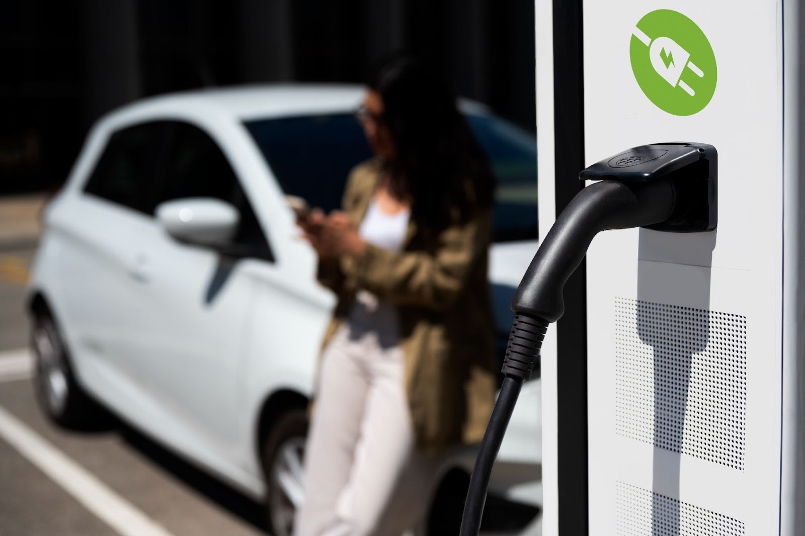 A foto em foco apresenta um ponto de recarga para carros elétricos, enquanto ao fundo, de forma desfocada, uma mulher está apoiada em seu carro segurando um celular.