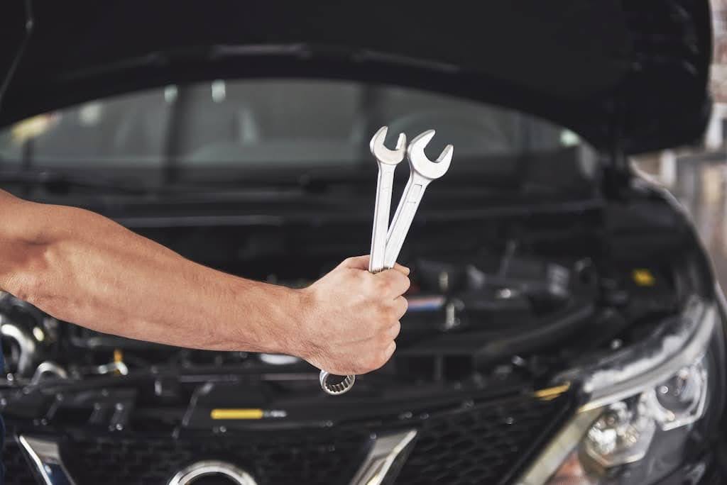 Foco no braço de mecânico segurando duas ferramentas com o capô aberto de um carro preto ao fundo.