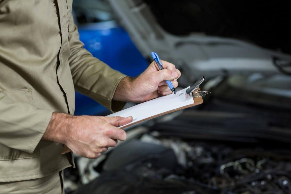 Foto mostra um mecânico segurando uma prancheta e fazendo anotações em frente a um carro com o motor exposto.