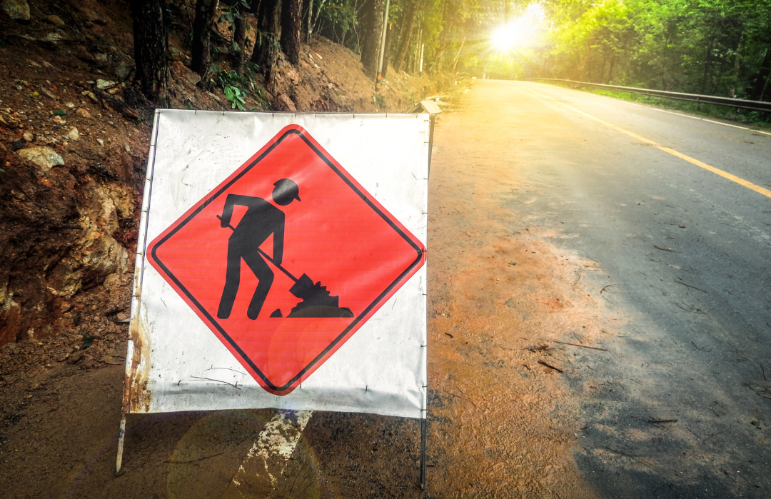 Foto mostra uma placa de sinalização de obras laranja à beira da estrada com desenho de um homem retirando terra com uma pá.