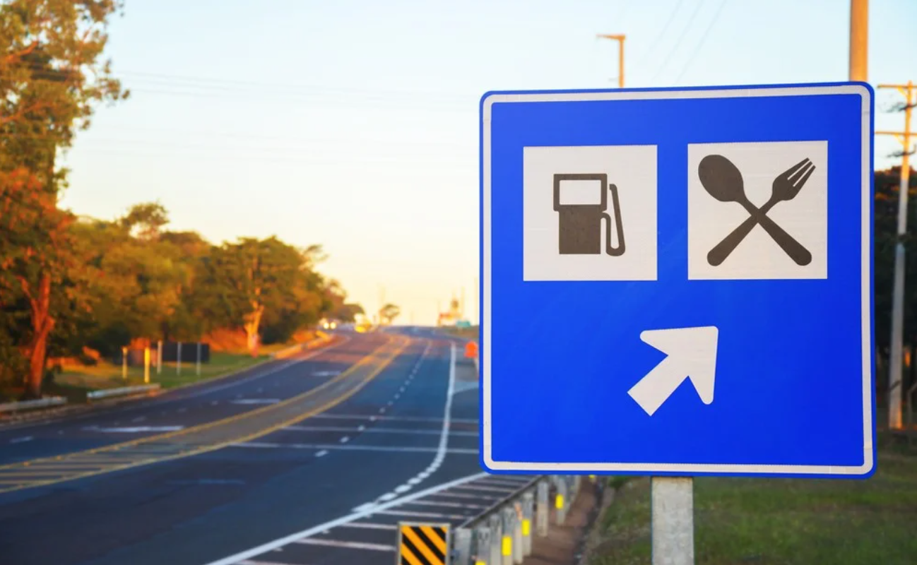 Foto mostra uma placa de sinalização de serviços auxiliares azul com o símbolo preto de uma bomba de gasolina e de um garfo e colher cruzados indicando restaurante.