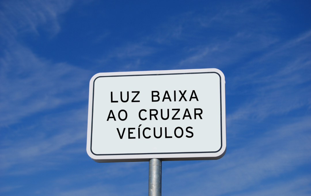 Foto mostra uma placa de sinalização educativa branca com letras pretas escrito “Luz baixa ao cruzar veículos”.