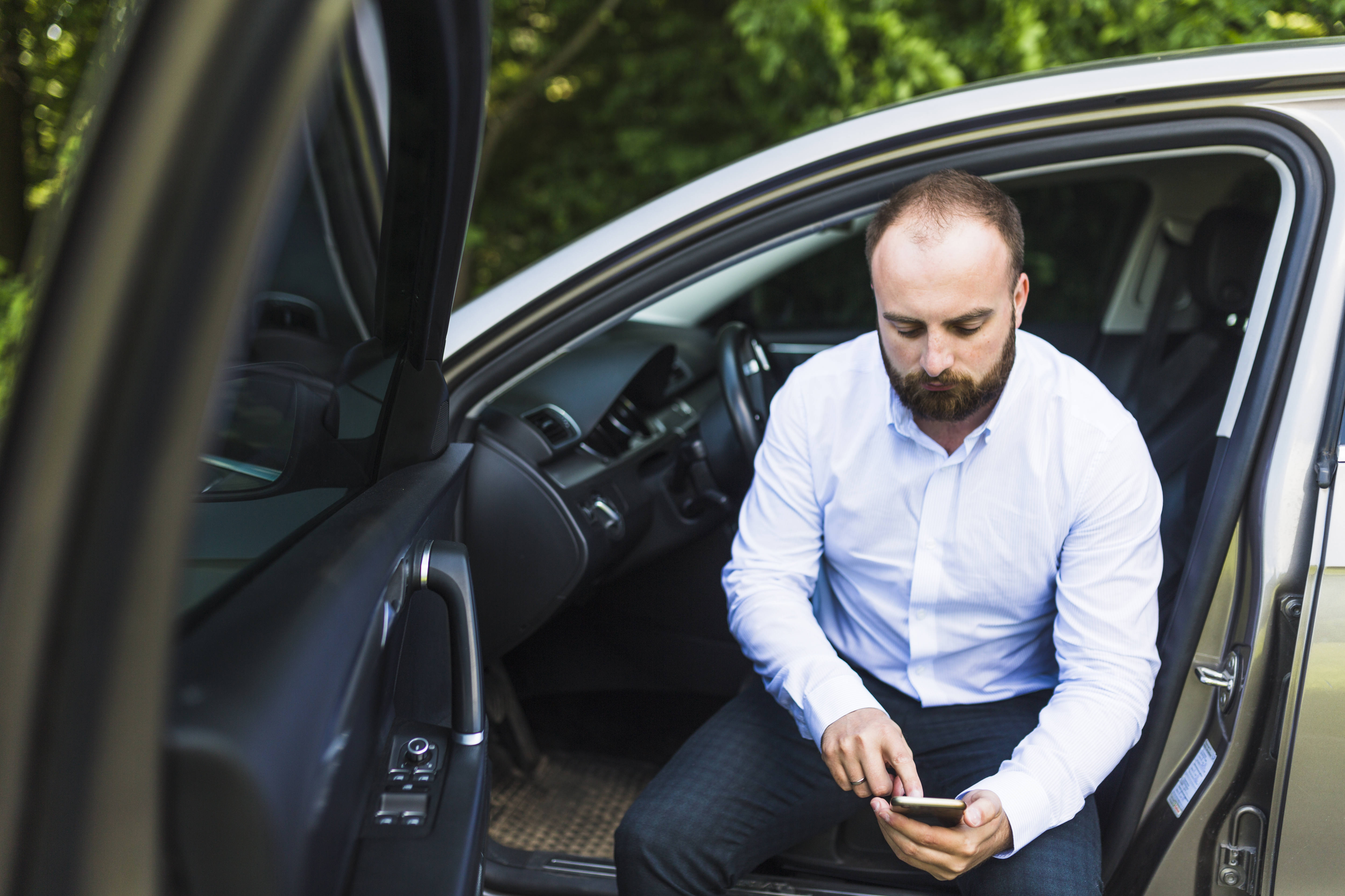 Homem de barba e camisa branca sentado na porta aberta de um carro, olhando para o celular que está segurando com as duas mãos. No fundo, há uma área externa com vegetação. A imagem representa a pesquisa da pergunta: o que é CRV e CRLV?