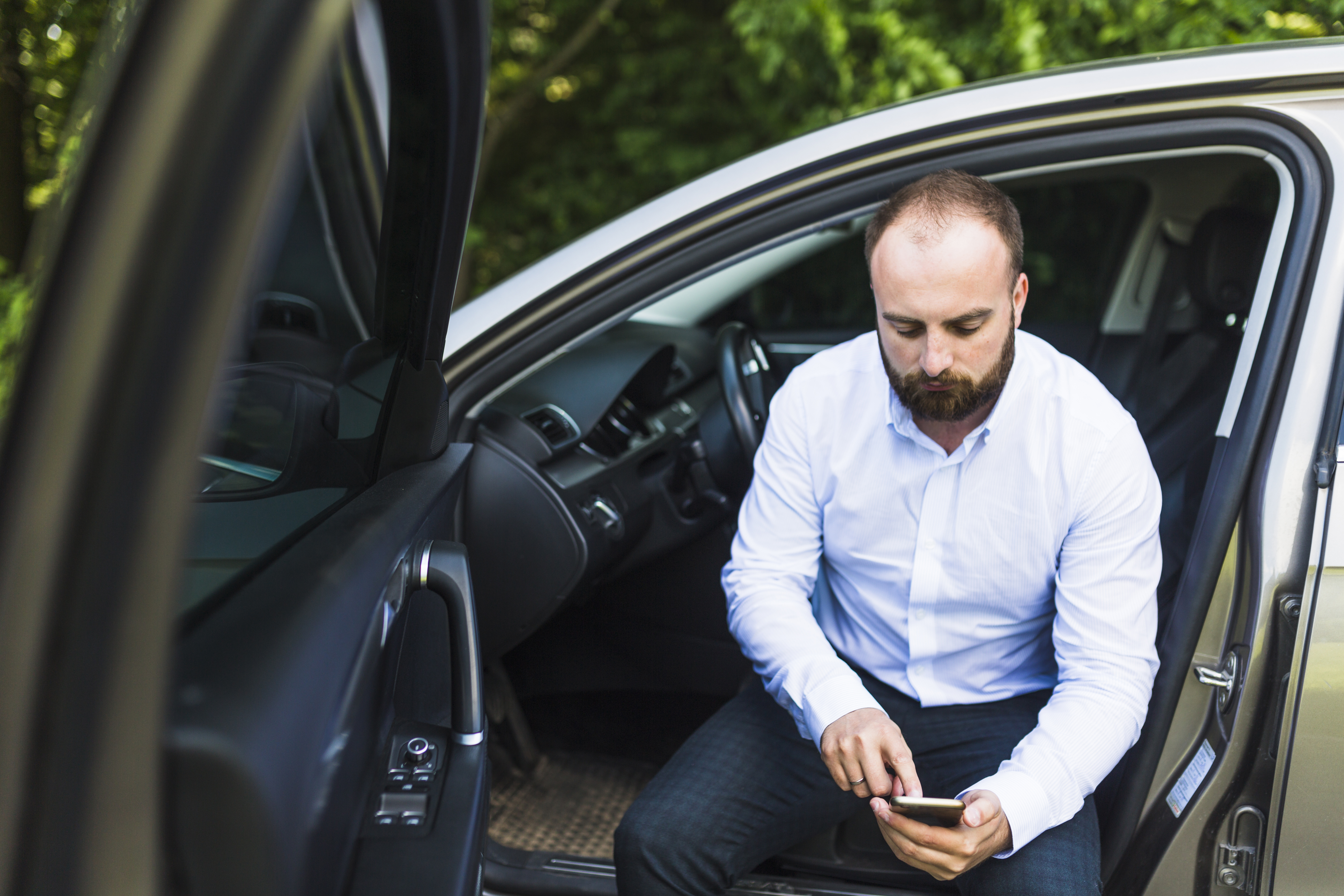 Homem de barba e camisa branca sentado na porta de um carro, olhando para o celular. Vegetação ao fundo.