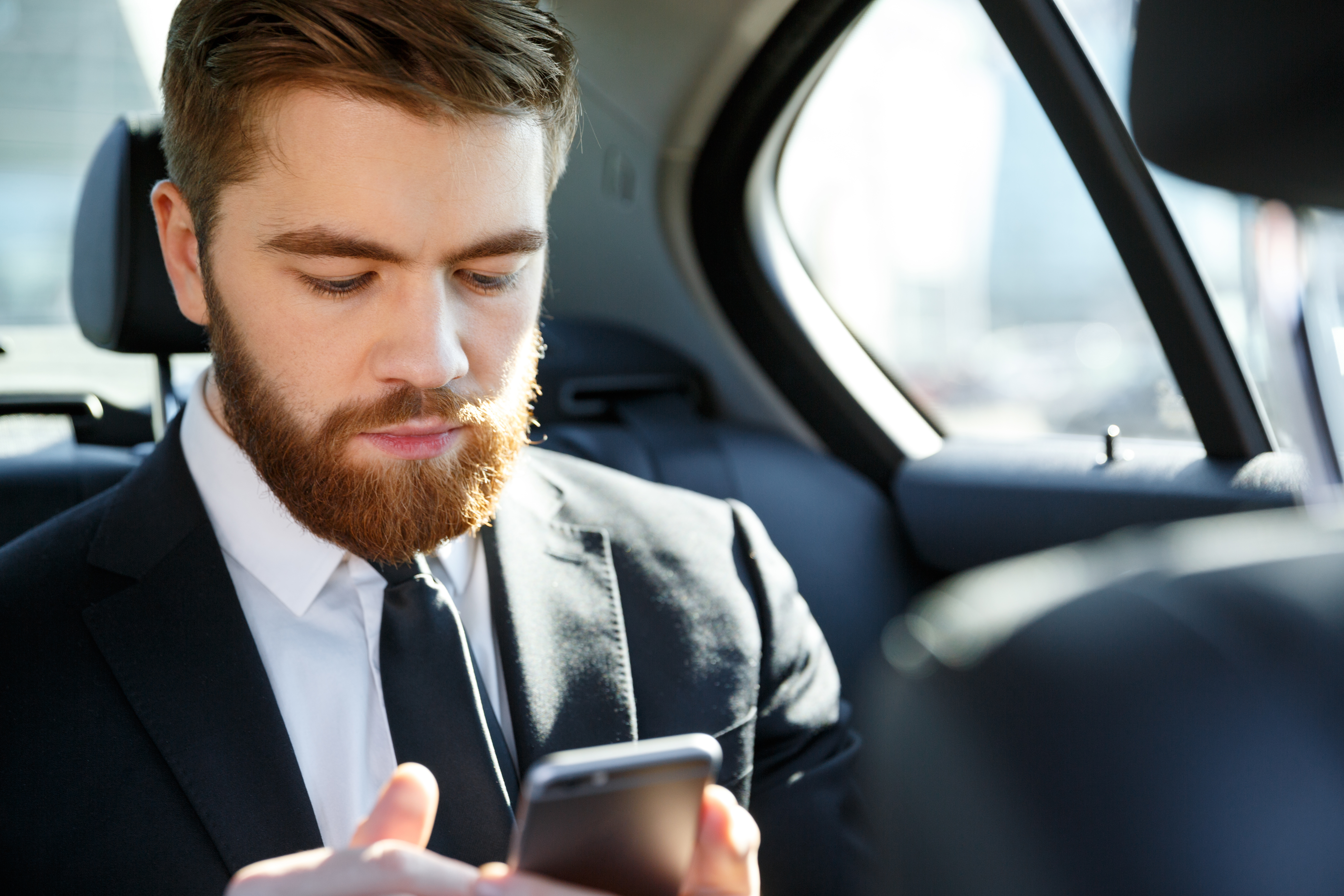 Homem de barba e terno sentado no banco traseiro de um carro, olhando para a tela de seu smartphone com uma expressão séria e concentrada.