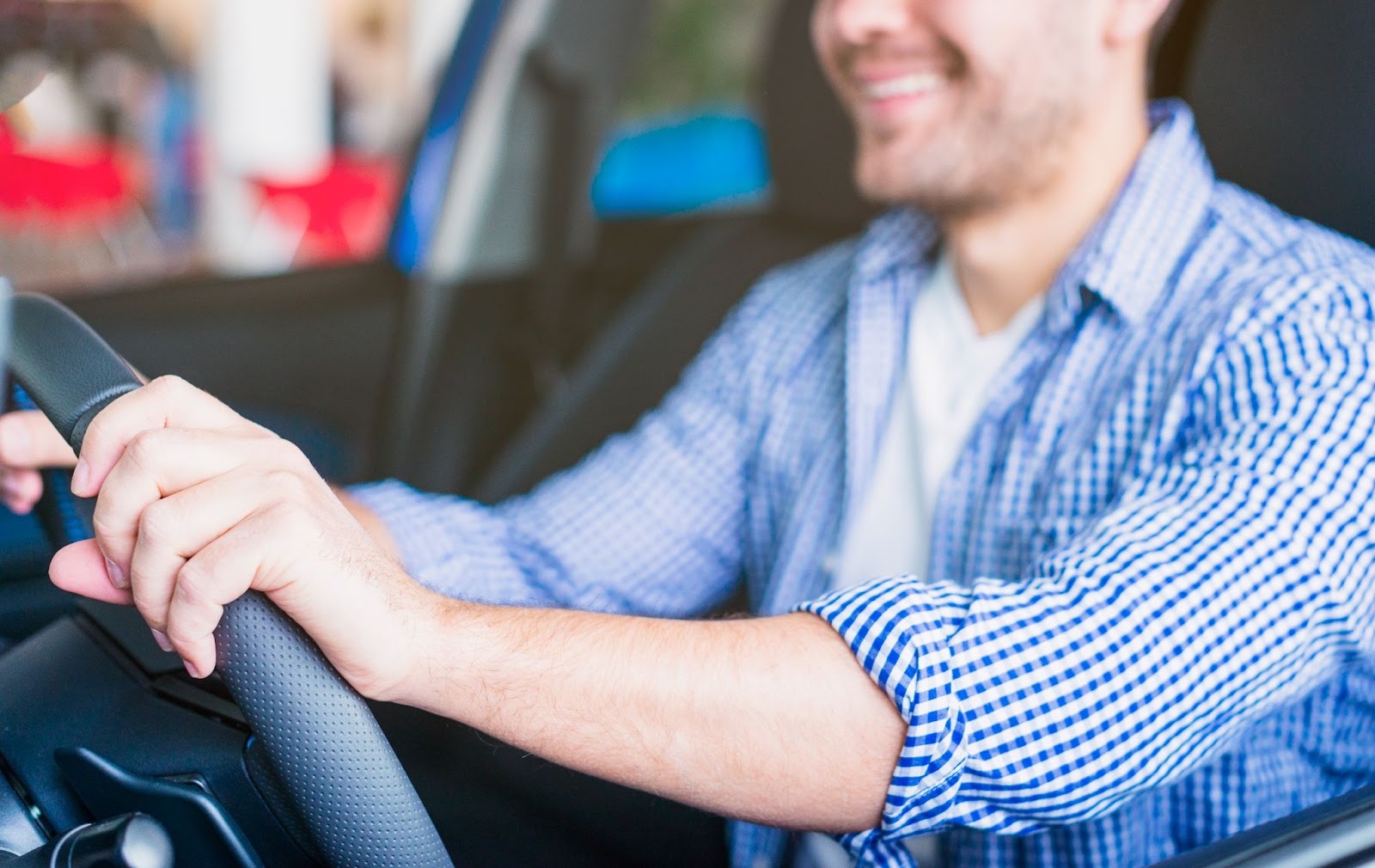 Homem motorista vestindo camisa xadrez azul está contente enquanto apoia as duas mãos no volante visando sua segurança no trânsito.