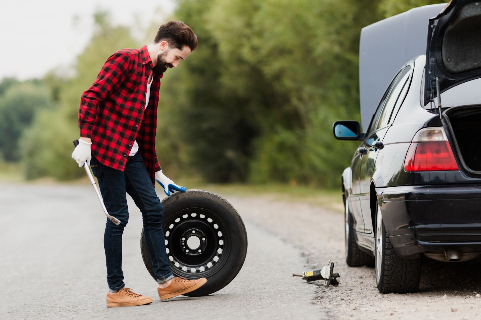 Homem vestindo camisa vermelha xadrez trocando pneu desgastado de seu carro preto na beira da estrada.