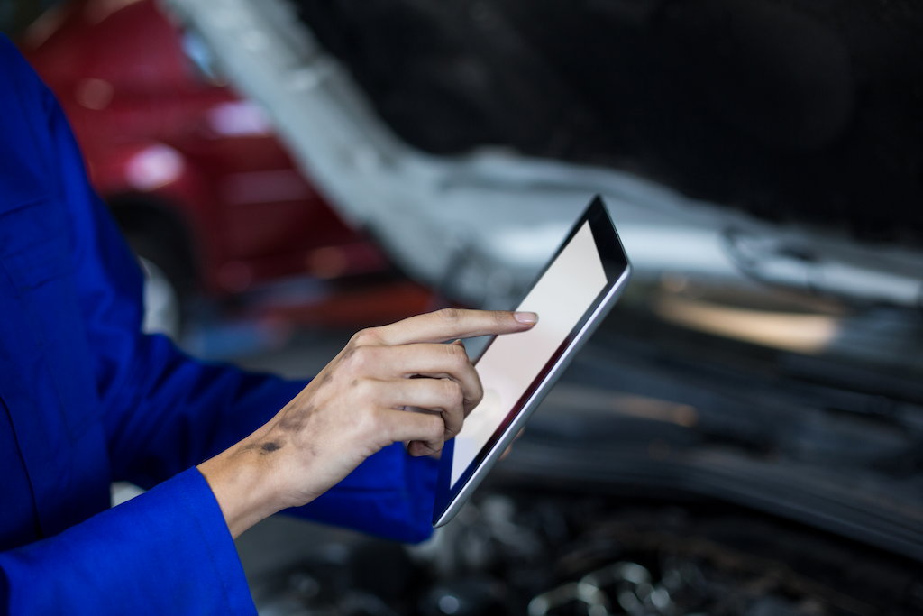 A imagem mostra as mãos de uma mulher mecânica em um macacão azul tocando um tablet na frente de um carro sendo analisado.