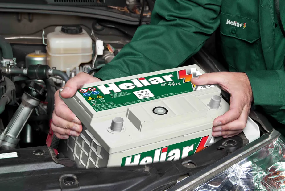 Imagem de uma pessoa, um mecânico de veículos, colocando uma bateria da marca Heliar em um carro. A pessoa está vestindo um macacão verde com a logo da empresa.