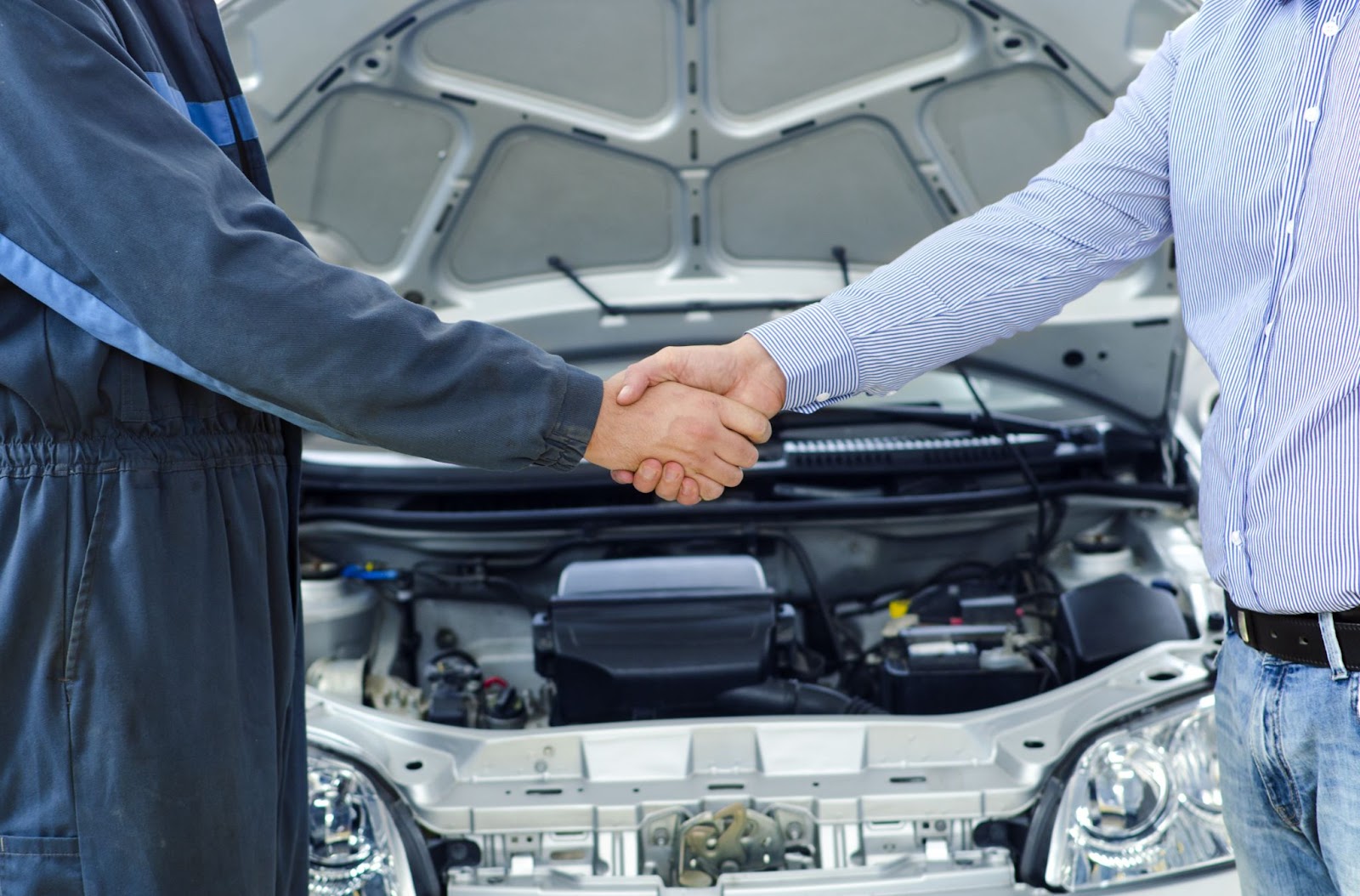 Imagem de um motorista e um mecânico em frente a um veículo com o capô aberto, cumprimentando-se com um aperto de mãos após o conserto do sistema de transmissão.