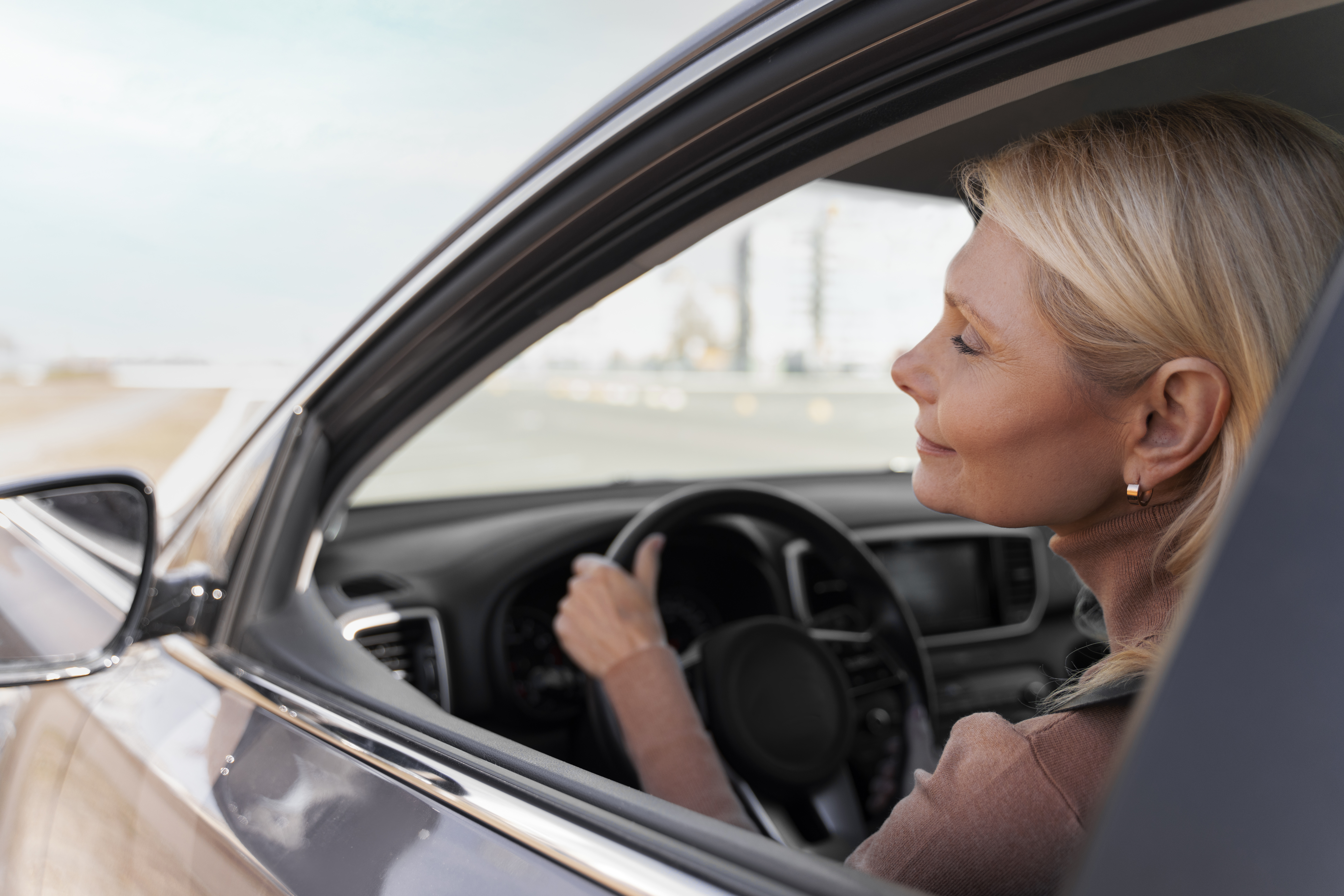 Imagem de uma mulher dirigindo um carro enquanto veste roupa social e apoia uma mão no volante.