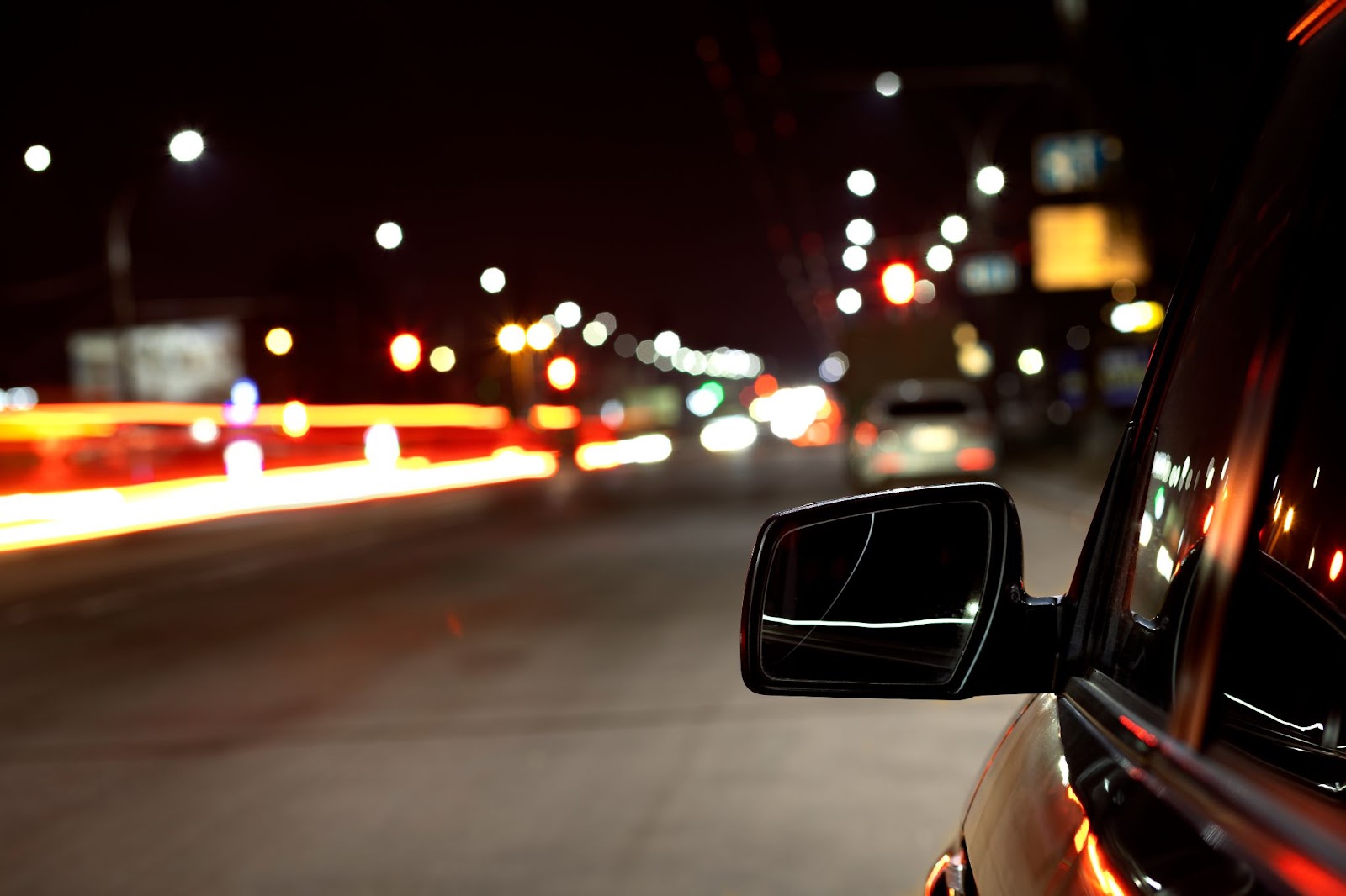 Imagem do retrovisor esquerdo em estrada à noite com luzes de farol de carro desfocadas ao fundo.