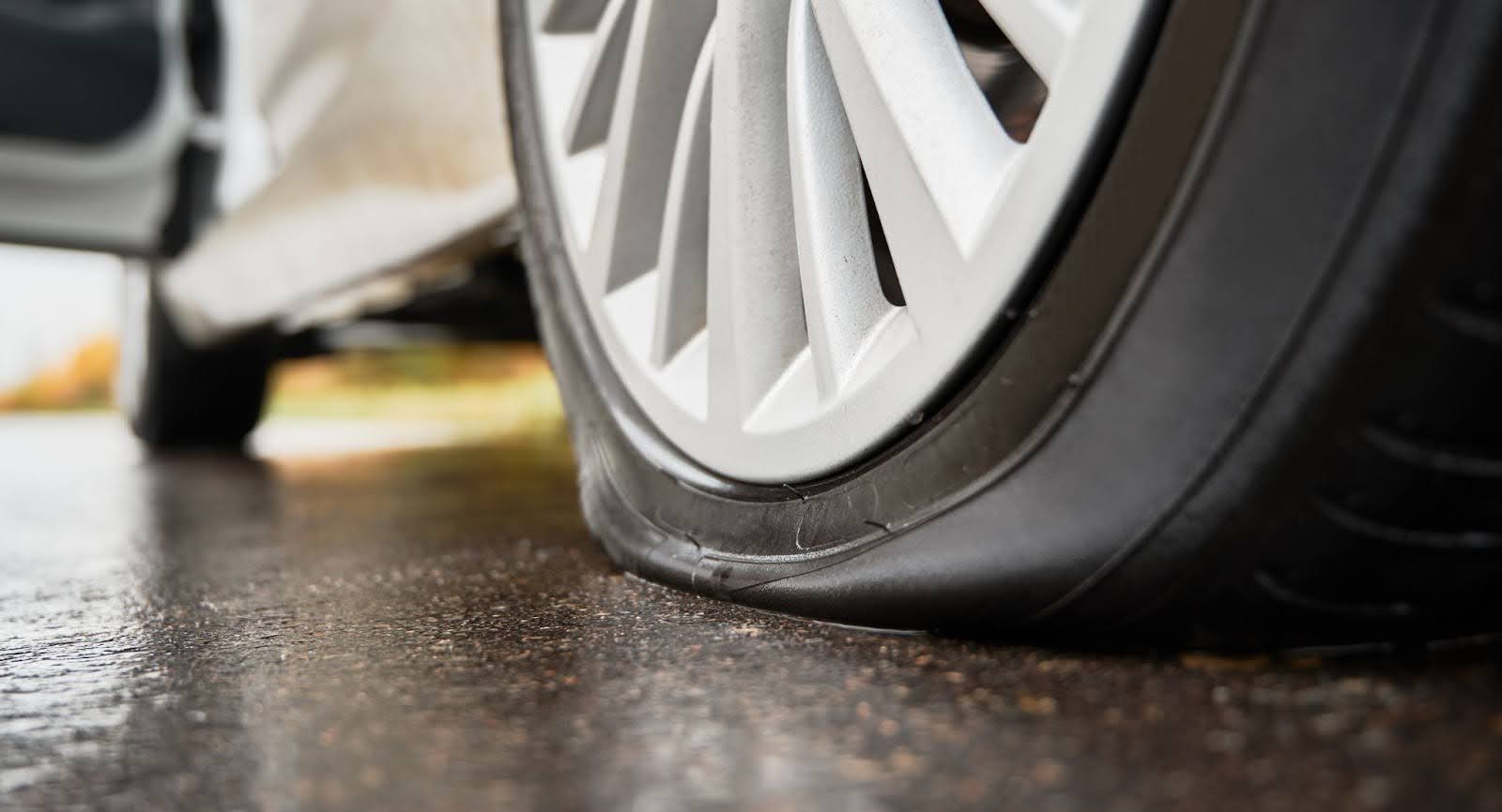 Imagem em close-up de uma roda de um carro branco com o pneu murcho sobre o asfalto.