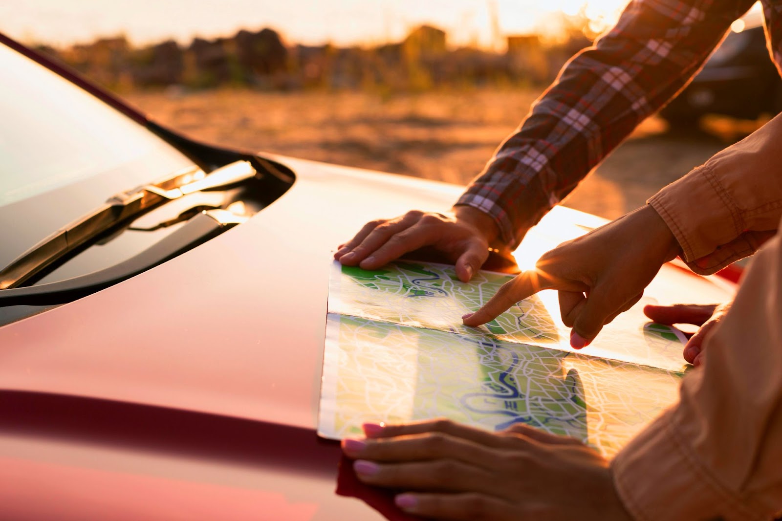 Imagem em close-up retrata duas pessoas apoiando um mapa sobre o capô de um veículo vermelho ao entardecer, planejando o itinerário de sua viagem de carro.