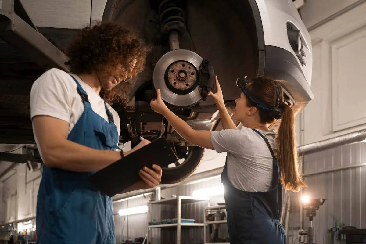 Imagem mostra dois mecânicos trabalhando no conserto de um carro. Um está segurando uma planilha e fazendo anotações enquanto a outra está tocando no disco e pastilha de freio.