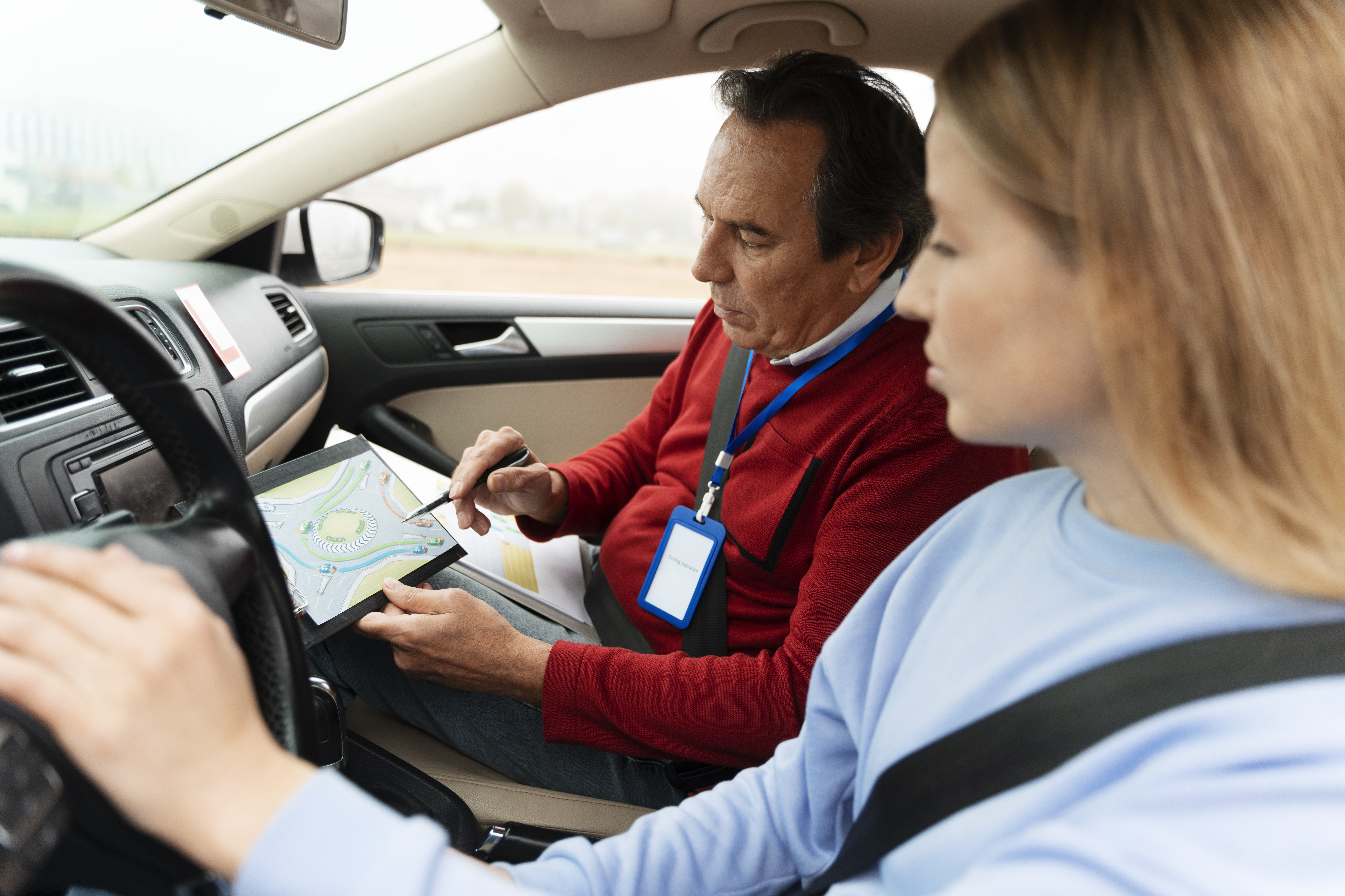 Imagem mostra um instrutor de autoescola e uma aluna, ambos dentro de um veículo. O instrutor está apontando para um mapa e explicando algo, enquanto a aluna observa.