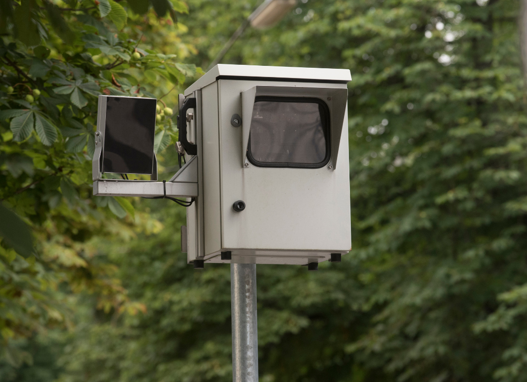 Imagem mostra um radar de velocidade cinza no topo de um poste de metal.