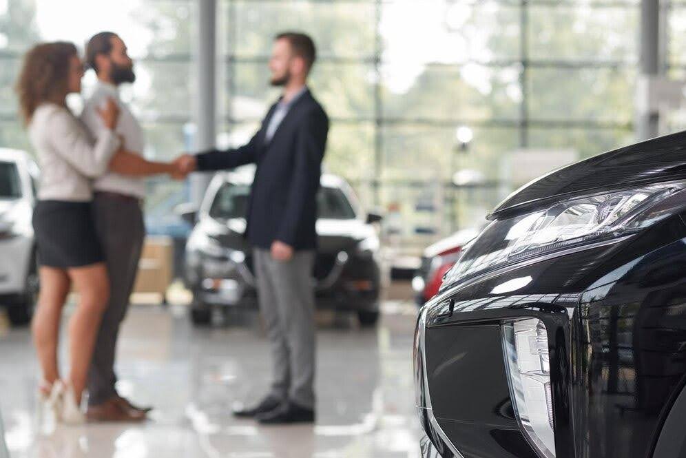 Imagem mostra uma mulher e um homem, que está apertando a mão de outro homem à sua frente. O ambiente é uma concessionária, representando a venda de um veículo.