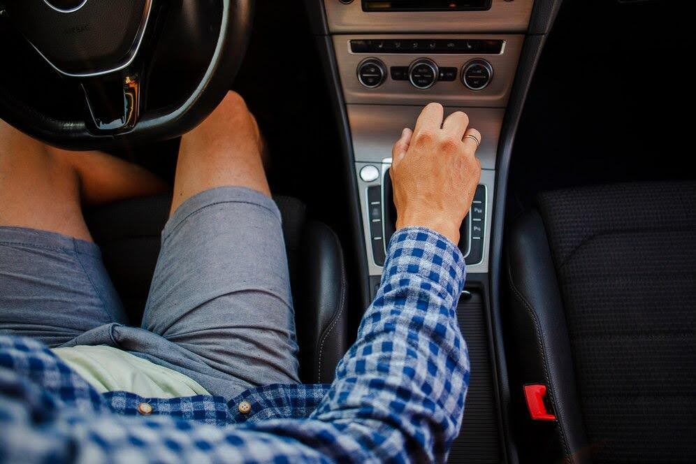 Imagem mostra a visão de cima e dentro de um carro de uma pessoa dirigindo um carro, com a mão na marcha.