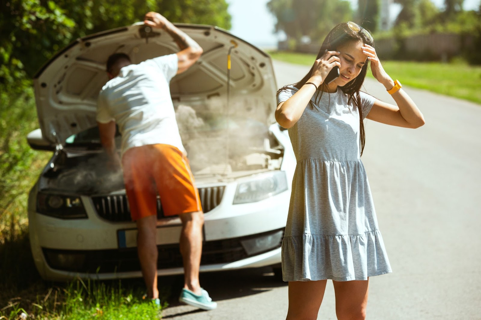 Uma mulher fala preocupada ao telefone na beira de uma estrada, ao fundo há um homem de bermuda laranja verificando o sensor de detonação de um motor que solta fumaça.