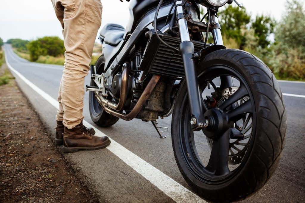 Motociclista de calça bege e bota marrom parado ao lado de uma moto preta no acostamento. Motociclista de calça bege e bota marrom parado ao lado de uma moto preta no acostamento.