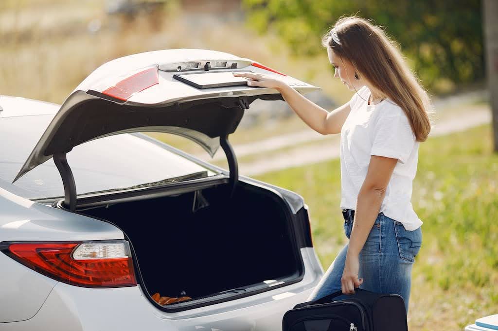 Mulher com camiseta branca e jeans segura uma mala preta enquanto abre o porta-malas de um carro prateado na rua. Mulher com camiseta branca e jeans segura uma mala preta enquanto abre o porta-malas de um carro prateado na rua.