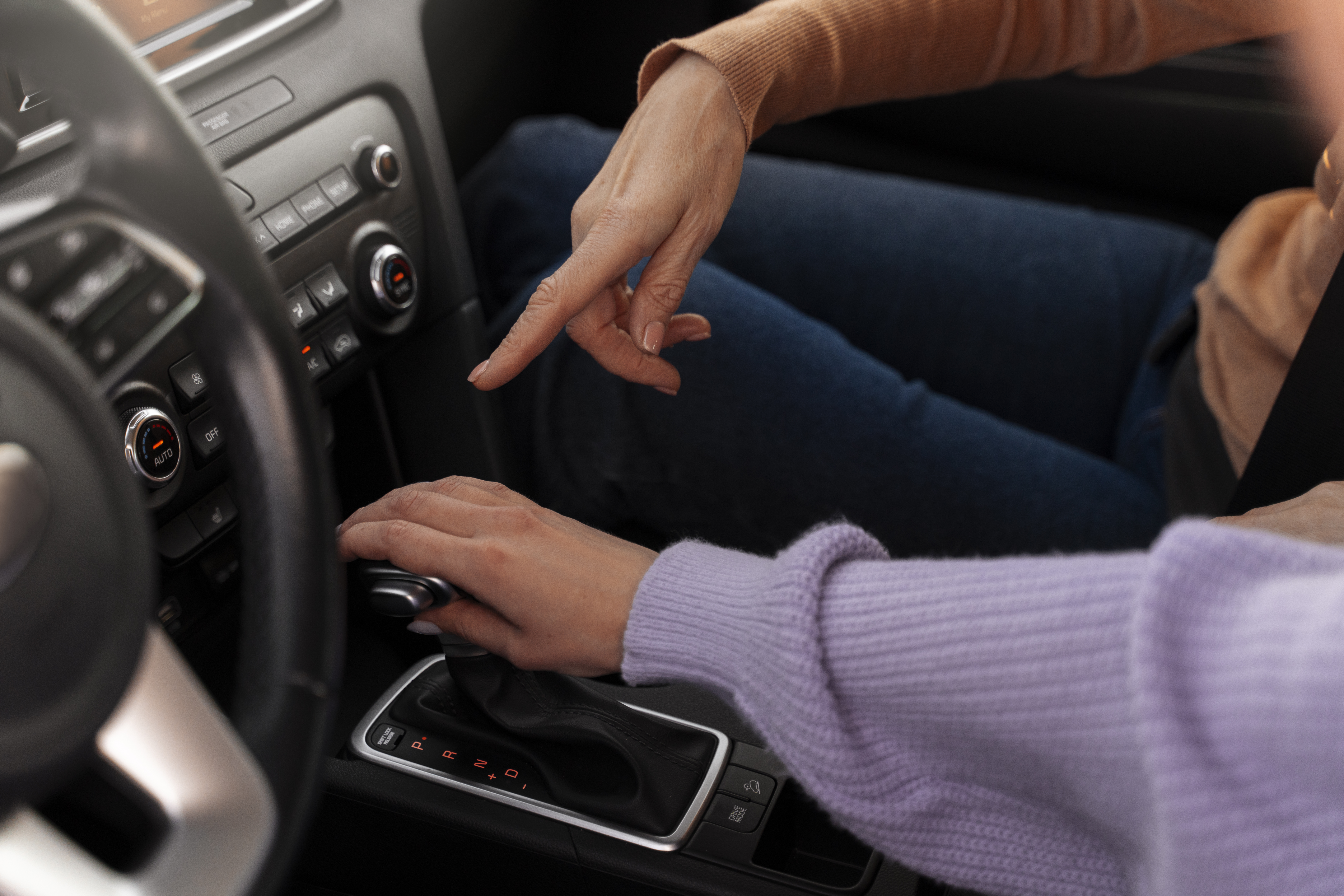 Mulher de blusa roxa sentada no banco do motorista segura o câmbio do veículo com a mão direita, enquanto outra mulher no banco do passageiro alerta sobre os cuidados com a correia dentada.