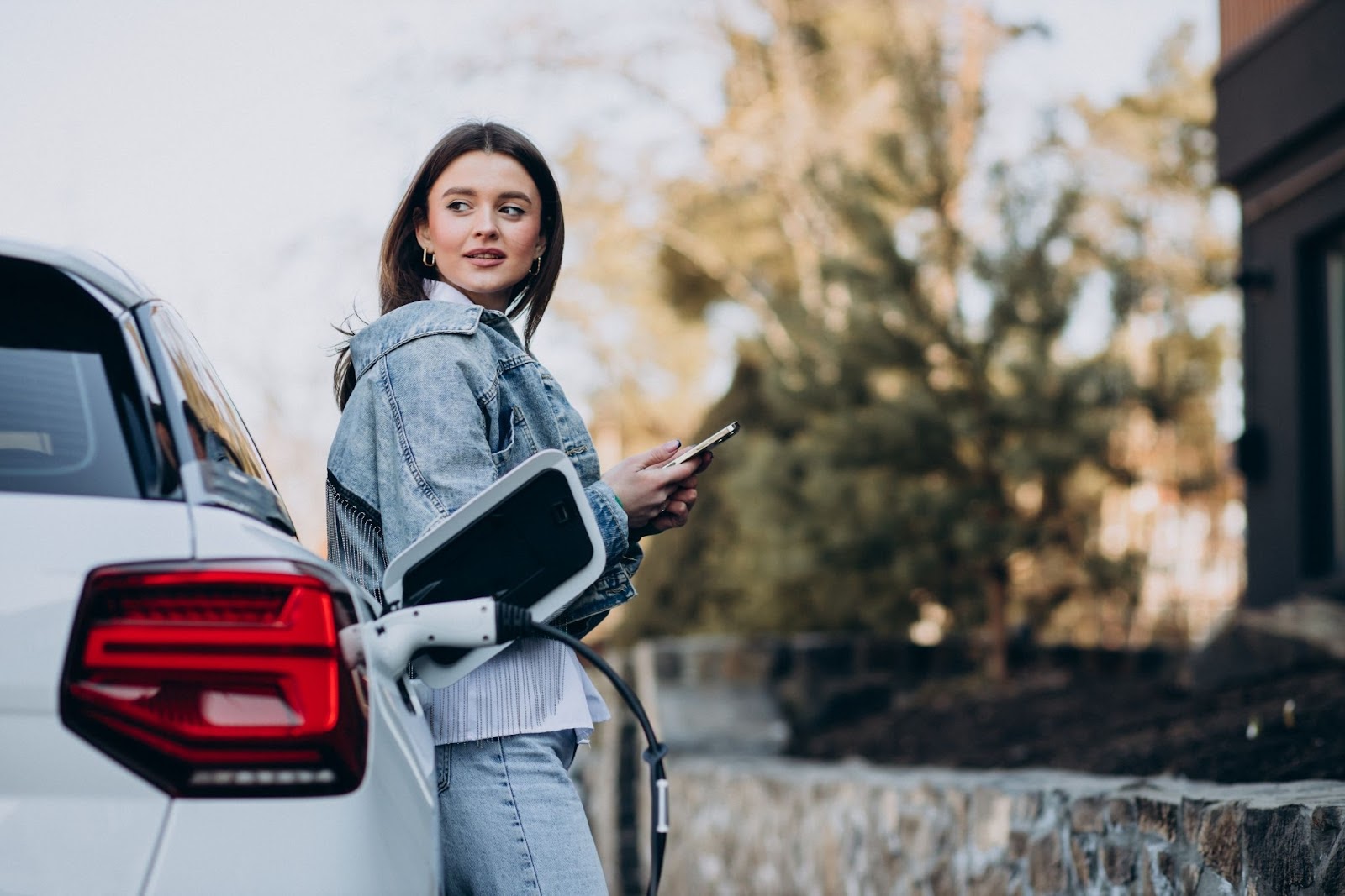 Mulher que sabe como funciona um carro elétrico, aguardando ao lado de seu veículo enquanto aguarda o processo de recarga. Ela está segurando um celular e olhando para o horizonte.