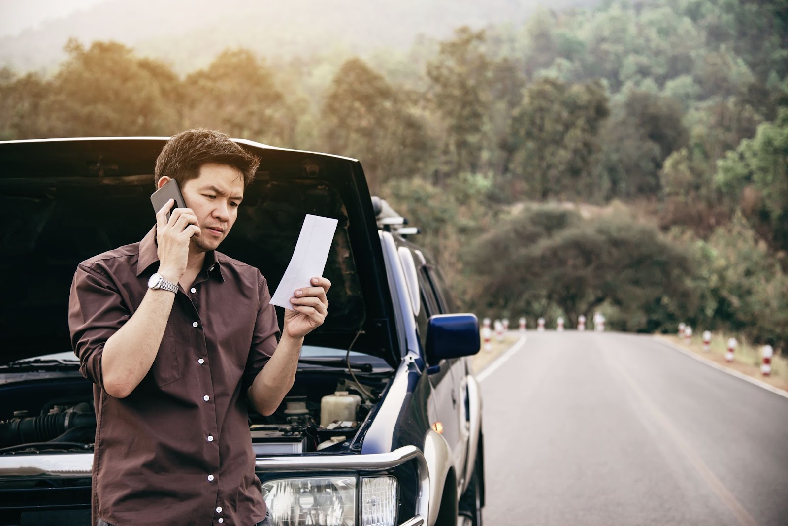 Na imagem há um homem de camisa marrom observando uma multa por falta de Certificado de Segurança Veicular enquanto fala ao telefone ao lado de um carro com o capô aberto.
