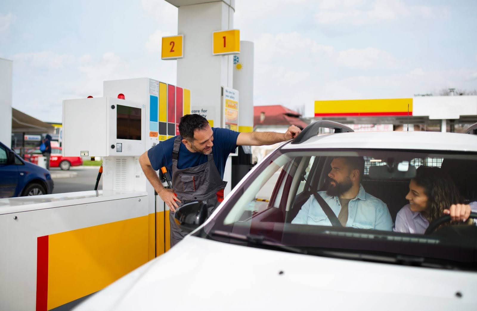 Na imagem um casal em um veículo branco é atendido por um frentista de macacão em um posto de gasolina que explica como calcular consumo de combustível.