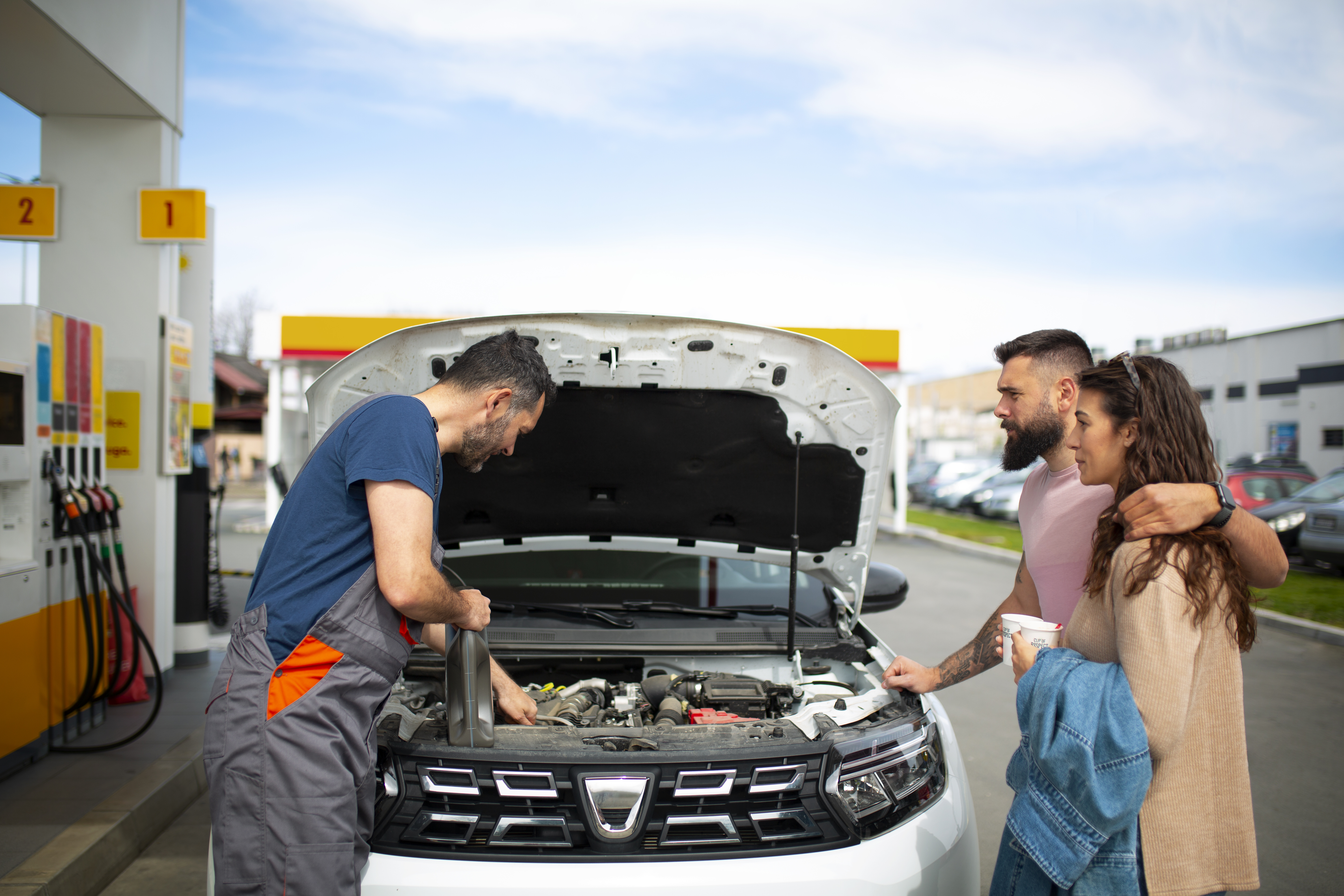 Na imagem um casal está parado ao lado de seu veículo branco com capô aberto enquanto um atendente do posto de gasolina verifica o motor e dá orientações sobre o filtro de combustível.