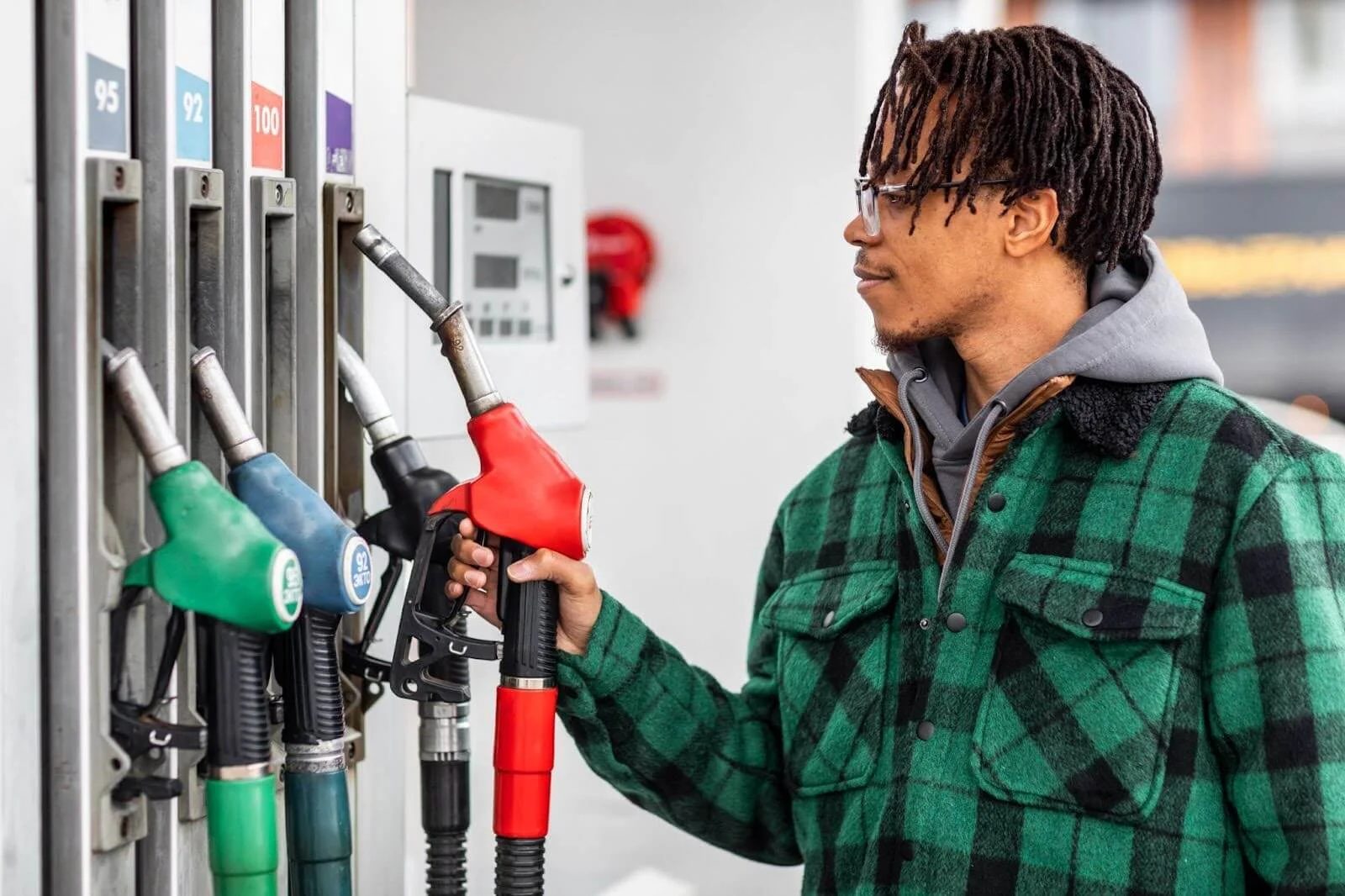 Na imagem um homem de casaco xadrez verde está em um posto de gasolina, segura o bico de abastecimento da bomba de gasolina enquanto se pergunta como calcular consumo de combustível.