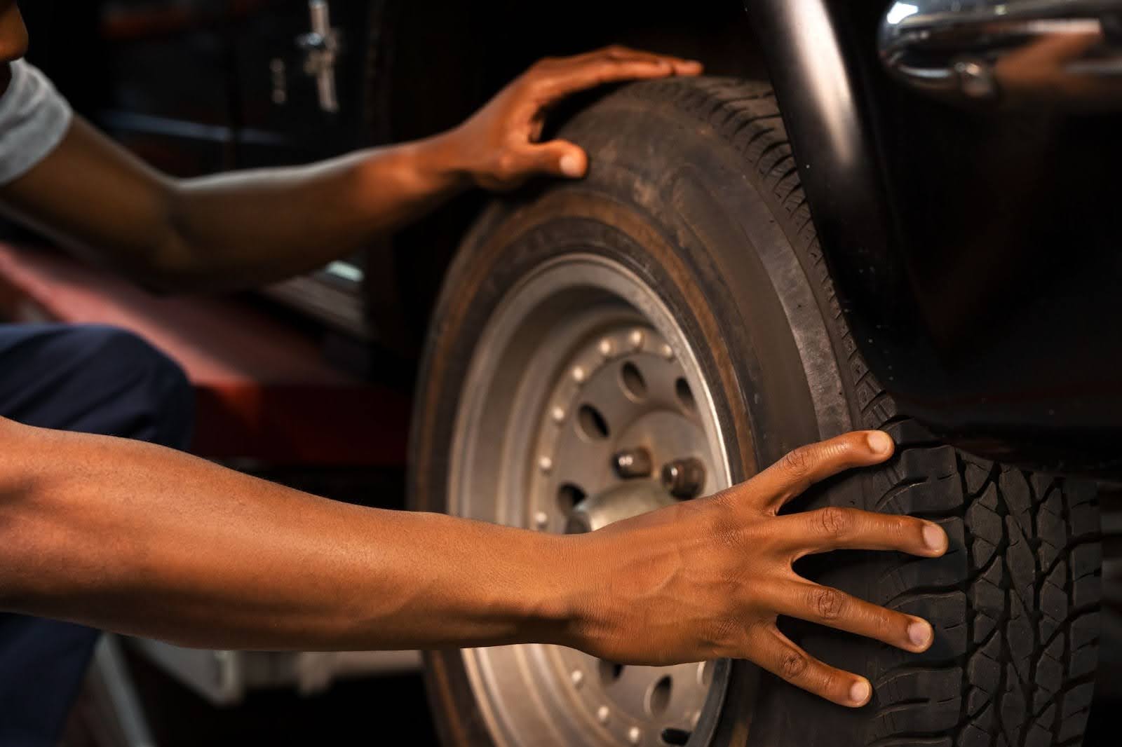 Na imagem um homem está abaixado com as mãos na roda de um carro preto, verificando se o pneu está furado.